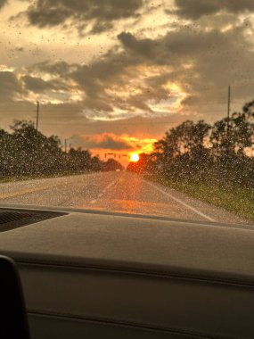 Gün batımında ıslak otoyolda giden bir arabanın içinde ön camda yağmur damlaları dramatik bulutlar ve ağaçlarla kaplı yolda parlayan ufuklar..