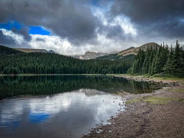 Sakin yansımalı dağ gölü, her yer yeşil orman kıyısı, dramatik fırtına bulutları ve mavi gökyüzü, ön planda kayalık plaj..