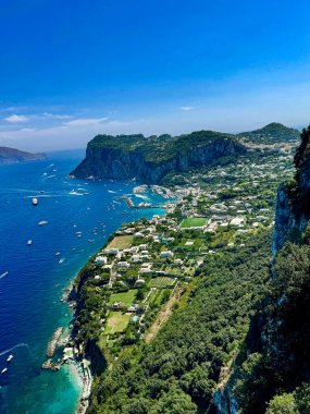 Uçurumları, turkuaz denizi, tekneleri ve berrak mavi gökyüzünün altında deniz kıyısında bir köyü olan yemyeşil bir yarımadanın havadan görünüşü.