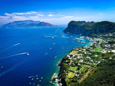Napoli Körfezi 'nin derin mavi sularına bakan Capri' nin dramatik uçurumlarının panoramik hava manzarası. Tekneler ve yatlar aşağıdaki denizi dolduruyor, yemyeşil teraslar ve berrak Akdeniz 'in altında engebeli kıyı şeridini kaplayan beyaz villalar var..