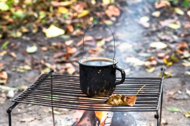 Metal black sooty mug on the grill on an open fire on the background of autumn leaves. Background.