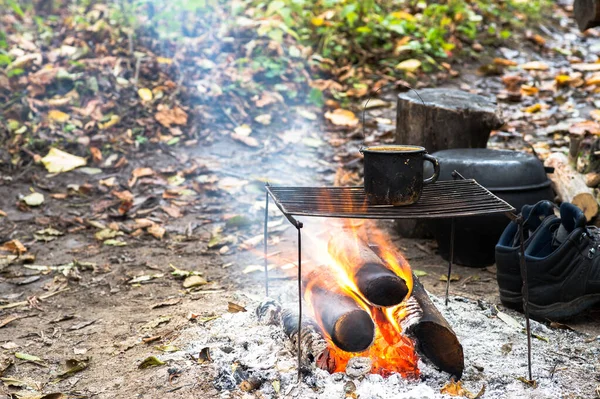 Metallschwarzer rußiger Becher auf dem Grill am offenen Feuer vor dem Hintergrund von Wanderausrüstung und Herbstlaub. — Stockfoto