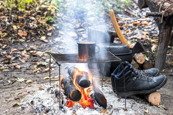 Metal black sooty mug on the grill on an open fire on the against the background of Hiking gear and autumn leaves.