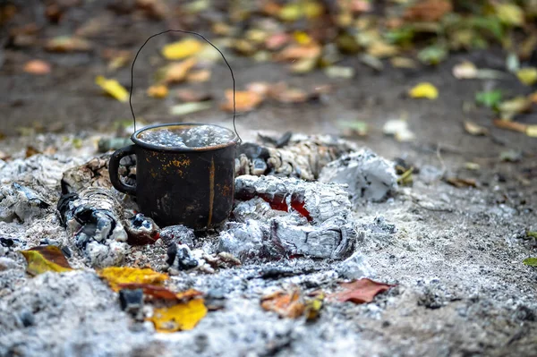 Metallschwarzer Räucherbecher mit kochendem Wasser auf den Kohlen eines Feuers auf dem Hintergrund von Herbstblättern. Hintergrund. — Stockfoto