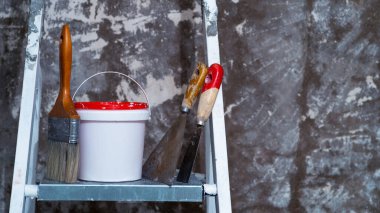 Bir fırça, kırmızı kapaklı bir kova ve spatula metal bir merdivenin üzerinde, üzerinde beyaz boya kalıntıları olan beton bir duvara karşı duruyor. Geçmişi. Onarım adımları kavramı.