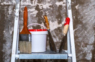 Bir fırça, kırmızı kapaklı bir kova ve spatula metal bir merdivenin üzerinde, üzerinde beyaz boya kalıntıları olan beton bir duvara karşı duruyor. Geçmişi. Onarım adımları kavramı.