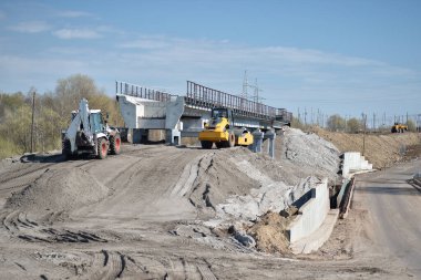 İnşaat ekipmanları yapay bir set oluşturur ve mavi yay gökyüzüne karşı karayolunun yanındaki yeni köprünün inşaat alanındaki zemini sıkıştırır. Arkaplan.