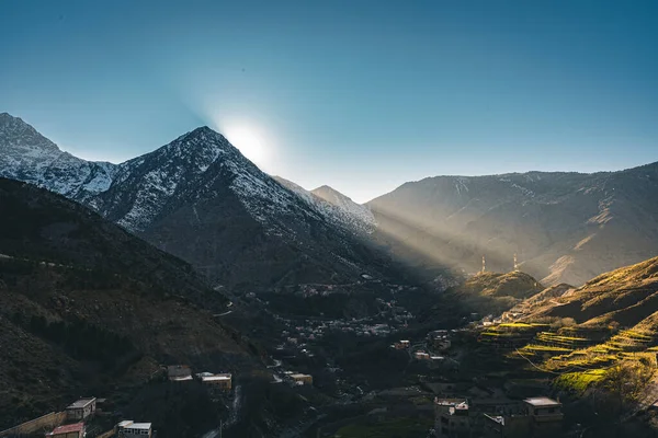 A view of sunset sunrise with sunstar in rural mountain village Imlil in High Atlas mountains Morocco in Africa. Snow covered peaks in background with lightrays and blue sky. — Stock Photo, Image