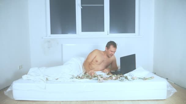Un uomo sdraiato su un materasso in camera, usa un computer portatile, si asciuga le lacrime con contanti — Video Stock