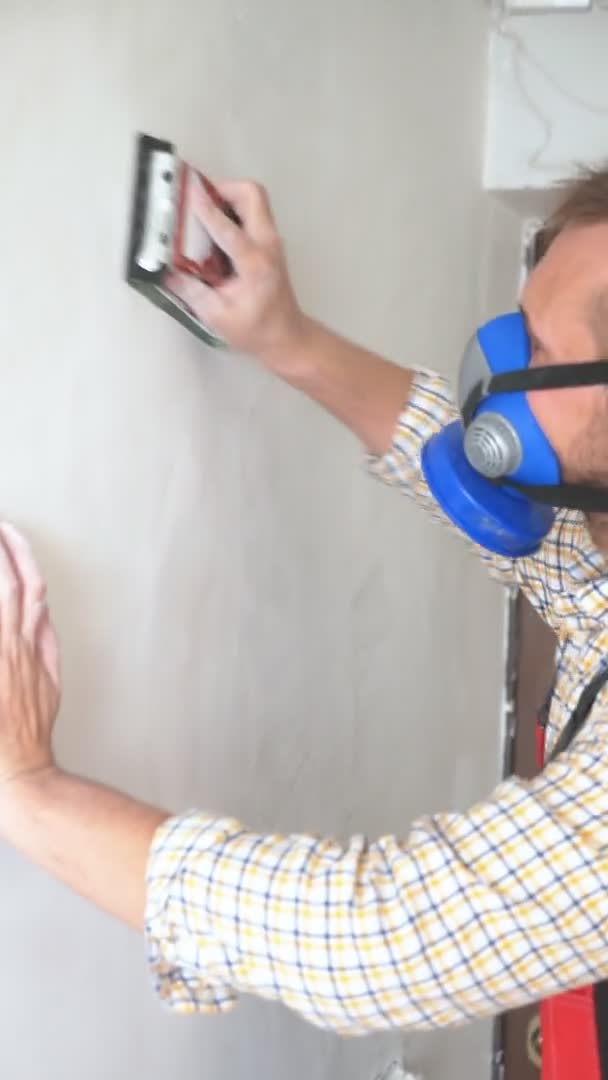 Verical. Un trabajador en un respirador está moliendo la pared con papel de lija — Vídeos de Stock