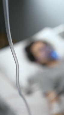 Arka plan bulanık. Hastane yatağında oksijen maskesi ve damlalığı olan bir adam var.