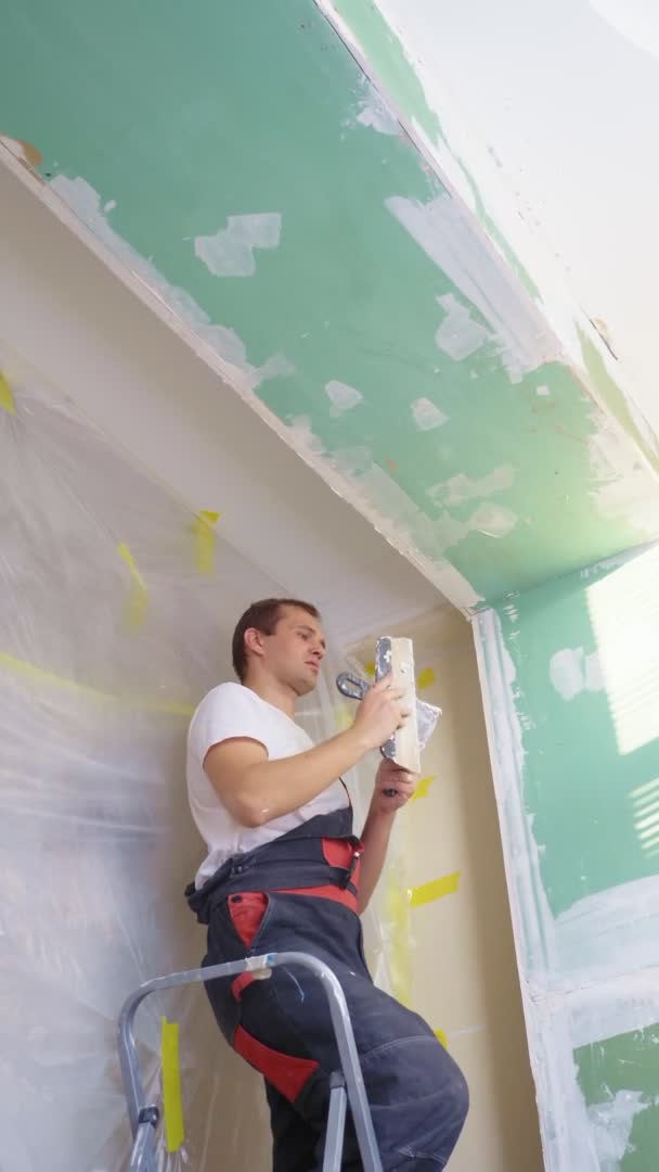 Verticale. un uomo in tuta applica intonaco con una spatola al muro — Video Stock