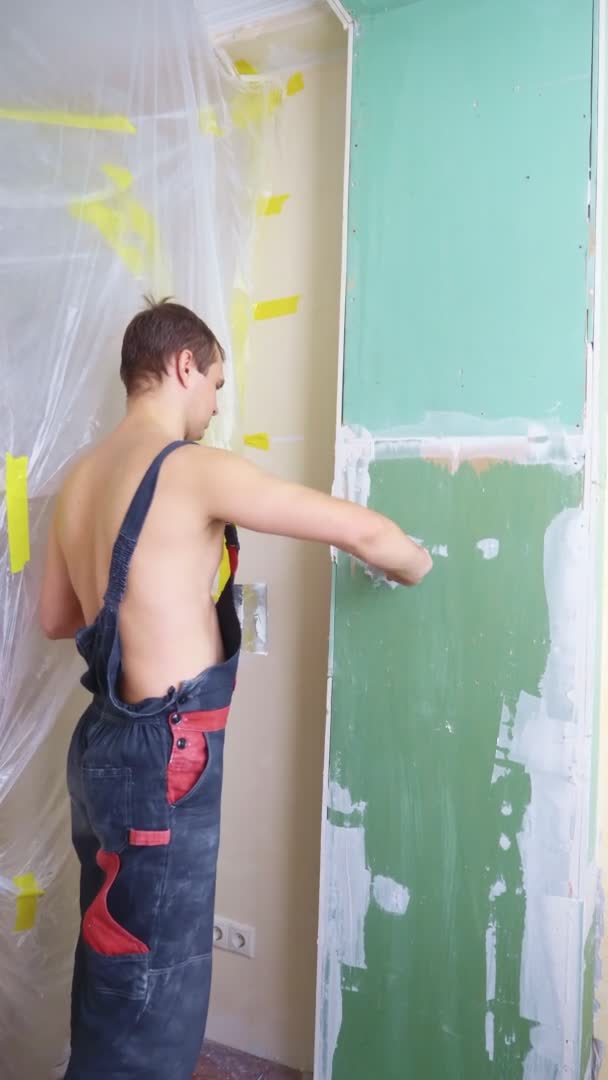 Verticaal. een man in een overall brengt pleister met een spatel aan op de muur — Stockvideo