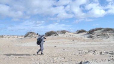 4K, profesyonel fotoğrafçı, yürüyüşçü, deniz kıyısının fotoğraflarını çekmek için tripod ve sırt çantasıyla çölde yürüyor..