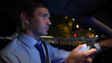 Arabadayken akıllı bir telefonla mesajlaşmak. Genç bir işadamı gece şehirde araba sürüyor ve akıllı telefonunu kullanıyor. 4k, ağır çekim.