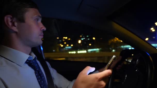 Mensajes de texto con un teléfono inteligente en el coche. un joven empresario está conduciendo un coche por la ciudad nocturna y utiliza su teléfono inteligente. 4k, cámara lenta — Vídeo de stock