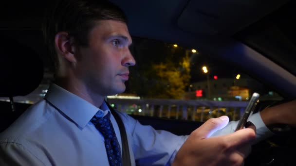 SMS avec un téléphone intelligent dans la voiture. un jeune homme d'affaires conduit une voiture dans la ville de nuit et utilise son smartphone. 4k, au ralenti — Video