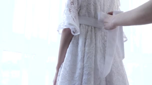 Ktoś zawiązał pasek na białej koronkowej sukience małej dziewczynki. 4k, strzelanie w zwolnionym tempie, promienie słoneczne — Wideo stockowe