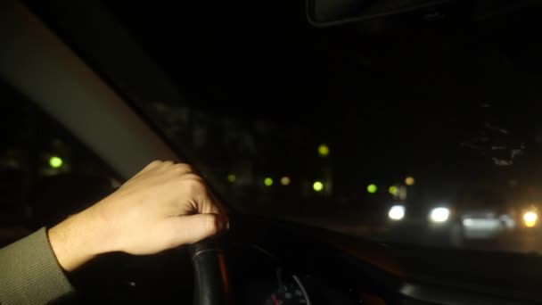 Main sur le volant de la voiture. Bonsoir, la nuit. Circulation à gauche. 4k. au ralenti, lumières floues devant la fenêtre. — Video