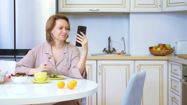 Mujer comiendo mientras está sentada en la cocina en la mesa y usando su teléfono inteligente — Foto de Stock