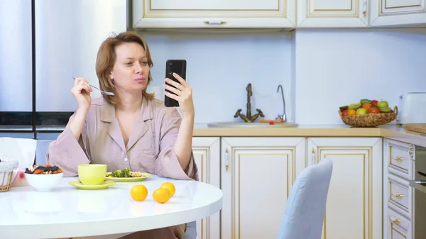 Mujer comiendo mientras está sentada en la cocina en la mesa y usando su teléfono inteligente — Foto de Stock