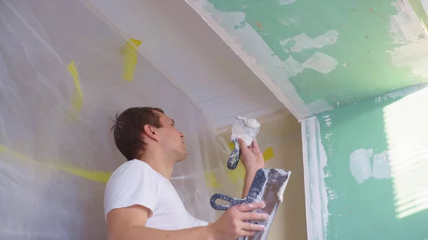 Un homme en salopette applique du plâtre avec une spatule au mur Image En Vente