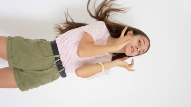 Vídeo vertical. chica adolescente delgada de pelo largo bailando alegremente sobre un fondo blanco — Vídeo de stock