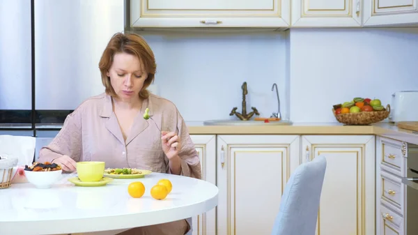 Wanita makan sambil duduk di dapur di meja di rumah Stok Gambar