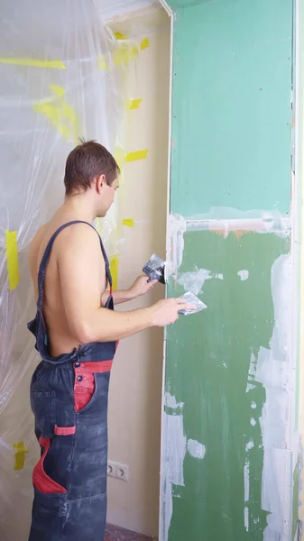 Un homme en salopette applique du plâtre avec une spatule au mur Photos De Stock Libres De Droits