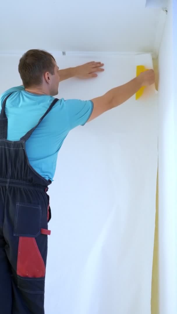 Video vertikal. seorang pria di overall glues wallpaper di dinding di apartemen — Stok Video