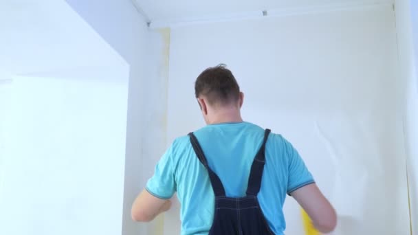 Мужчина в комбинезоне клеит обои на стенах в квартире — стоковое видео