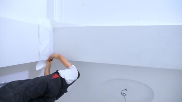 Vídeo vertical. un hombre joven en overoles pega papel pintado al techo — Vídeos de Stock