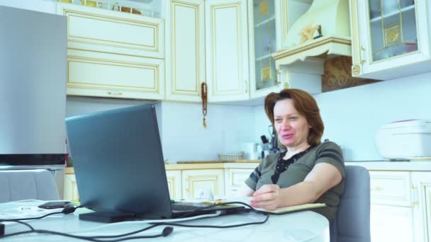 Женщина принимает участие в видеоконференции на ноутбуке, сидя дома на кухне — стоковое видео