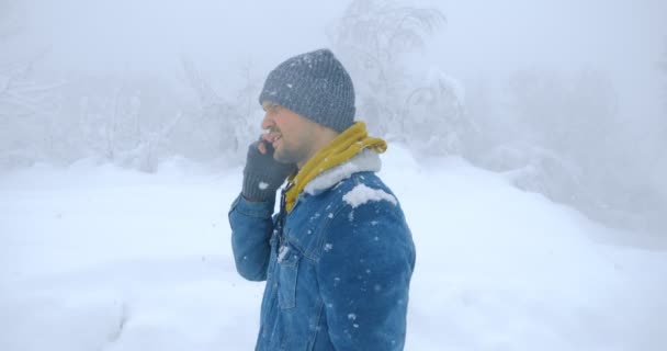Um homem falando em um smartphone andando em um parque de inverno durante uma queda de neve — Vídeo de Stock