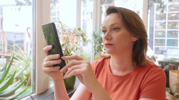 Mujer en una camiseta escarlata utiliza un teléfono inteligente mientras está sentado en un café — Vídeos de Stock