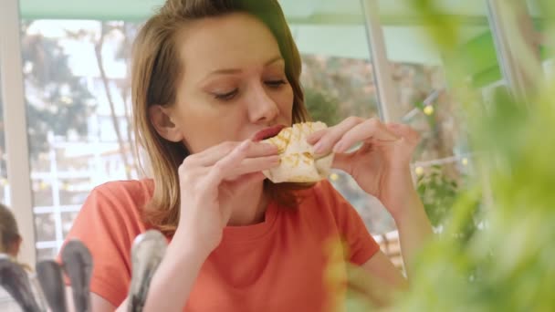 Retrato de uma mulher em uma camiseta escarlate comendo shawarma enquanto sentado em um café pela janela — Vídeo de Stock