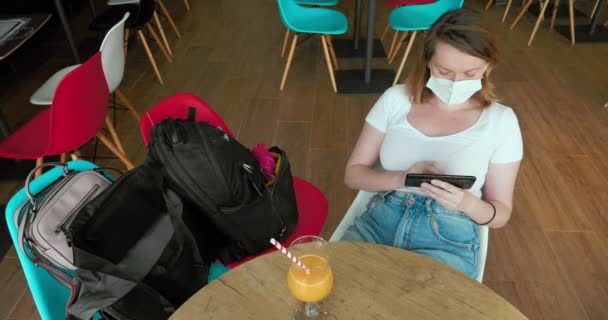 戴着面具的女游客坐在空荡荡的咖啡店里，用智能手机喝橙汁 — 图库视频影像