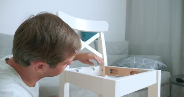 Ein Mann in Heimkleidung baut einen Stuhl mit einem Schraubenzieher selbst zusammen — Stockvideo