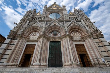 İtalya 'daki Siena Katedrali' nin Gotik cephesi. Karmaşık polikrom mermer, detaylı heykel elementleri ve mavi parçalı bulutlu gökyüzünün altında merkezi bir gül penceresi..
