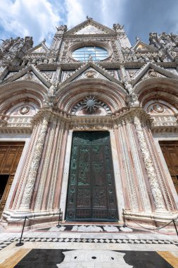 Karmaşık mermer kaplamalı, bronz kapılı ve detaylı frizlerle Siena Katedrali 'nin Gotik cephesi. Bulanık gökyüzüne karşı dramatik bir yükseliş perspektifi. Siena, İtalya 'da yer almaktadır.