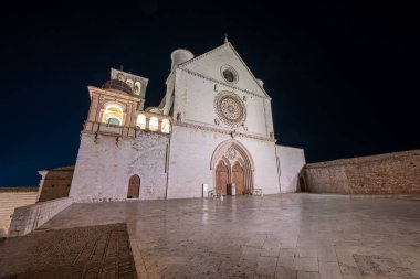 İtalya 'nın Assisi kentindeki Papalık Bazilikası, gece gökyüzüne karşı aydınlatılmış çarpıcı bir beyaz taş cephe ve karmaşık gül penceresiyle Romanesk ve Gotik mimarisini sergiliyor..
