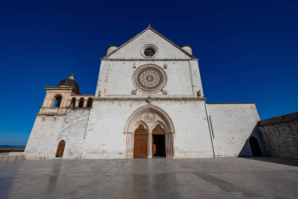 The Papal Basilica of Saint Francis in Assisi features Gothic architecture with a striking rose window. Its white limestone facade contrasts against the deep blue sky. Located in Assisi, Italy.