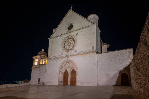 Gothic basilica in Assisi, Italy, famous for 13th-century architecture, features rose window, intricate facade, illuminated at night, site linked to Saint Francis, important pilgrimage destination.