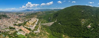 Rocca Albornoziana, bir 14. yüzyıl kalesi ve İtalya 'nın Spoleto kentindeki Ponte delle Torri su kemeri. Kireçtaşı yapılar açık mavi gökyüzünün altındaki yemyeşil tepelerin ortasında.