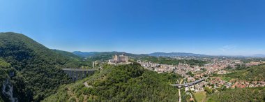 Ortaçağ Rocca Albornoziana kalesi, 14. yüzyıl Ponte delle Torri su kemeri yanındaki taş duvarlar ve kulelerle ufuk çizgisine hakim. Spoleto üzerinde açık bir yaz gününde hava manzarası.