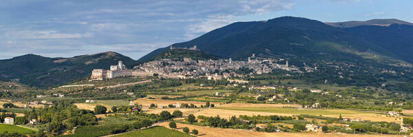 Aerial view of the Basilica of San Francesco in Assisi, Italy. The medieval structure with its distinctive stone facade sits against verdant hills under a cerulean sky, embodying spiritual heritage.