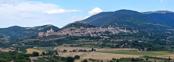 Aerial view of the Basilica of San Francesco in Assisi, Italy. The medieval structure with its distinctive stone facade sits against verdant hills under a cerulean sky, embodying spiritual heritage.