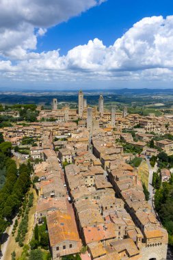 San Gimignanos ortaçağ taş kuleleri ve Tuscan kırsal kesiminin tepelerinde yaz bulutları altında yükselen terasotta çatıları..