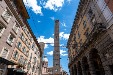 Bologna 'daki ortaçağ Asinelli Kulesi' nin geniş sokak manzarası, Porticoed Palazzi ve yaz gökyüzü altında modern dükkanlar tarafından çevrelenmiş tuğla yapı.