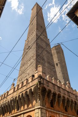 Bologna 'daki ortaçağ Asinelli ve Garisenda tuğla kulelerinin alçak açılı görüntüsü, yazlık gökyüzünün altındaki saray ve tramvay kablolarının üzerinde yükseliyor..